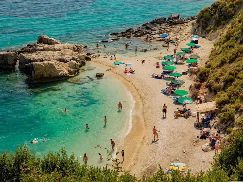 People swim in clear waters that are dotted with green parasols and backed by a forest.