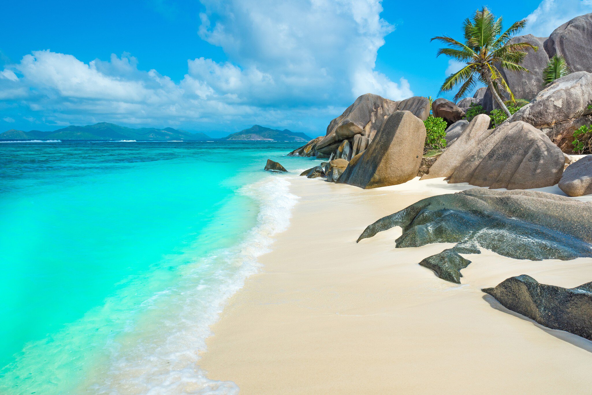 Smooth grey granite boulders and a leaning palm above pale sand, with shallow turquoise water and green islands on the horizon.
