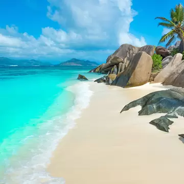 Smooth grey granite boulders and a leaning palm above pale sand, with shallow turquoise water and green islands on the horizon.