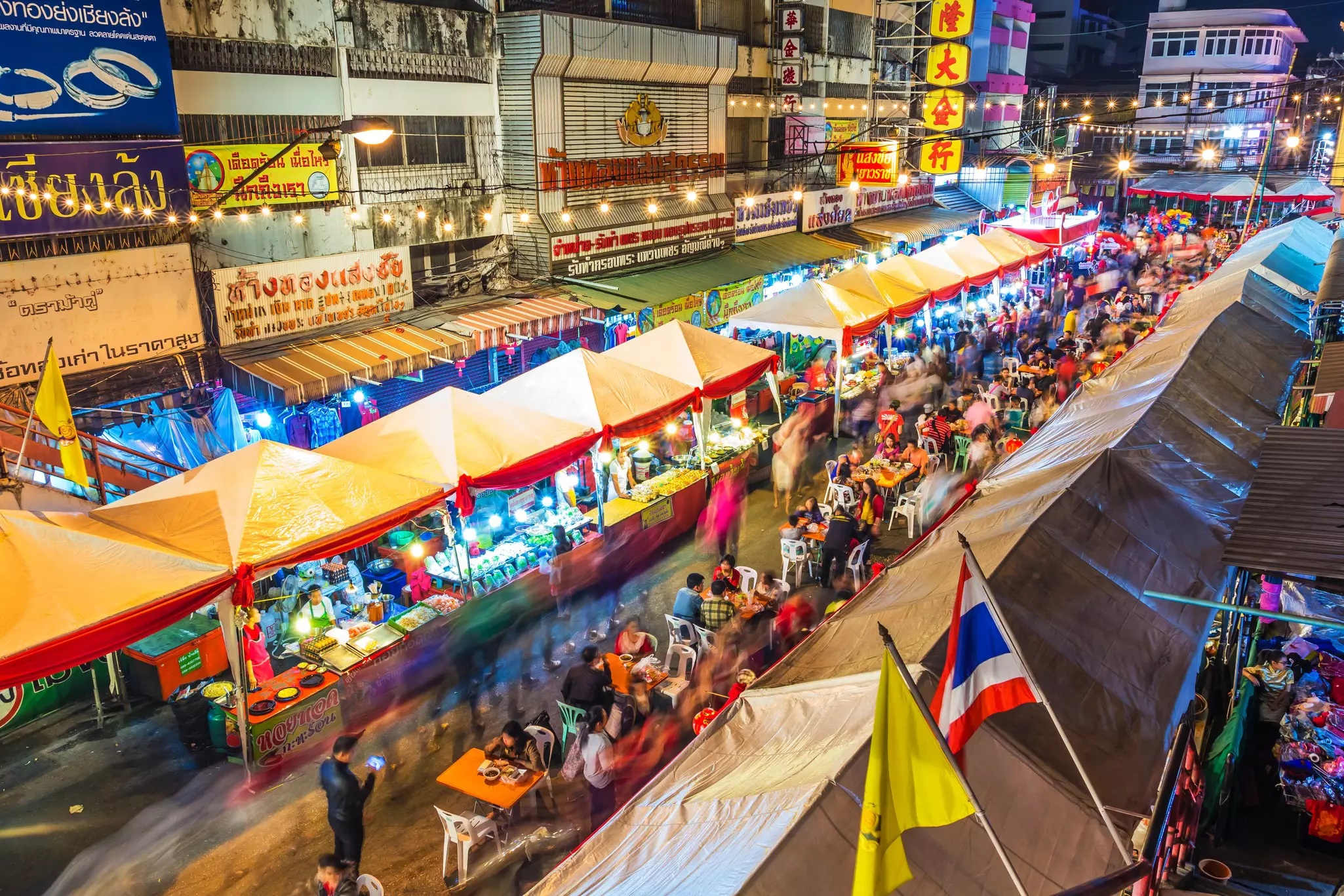 A long street lined with vendors with dining tables set out in the center. Many people shop and eat at the night market.