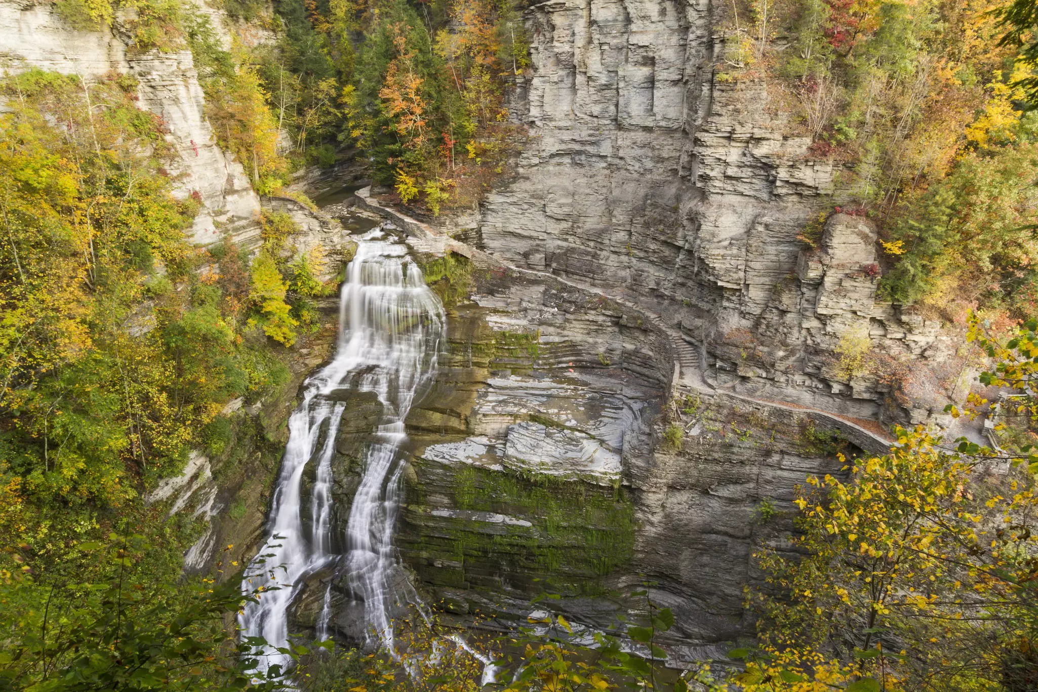 Aerial view of waterfall cascading down gray cliffs with autumnal-colored trees surrounding it on an overcast day.
