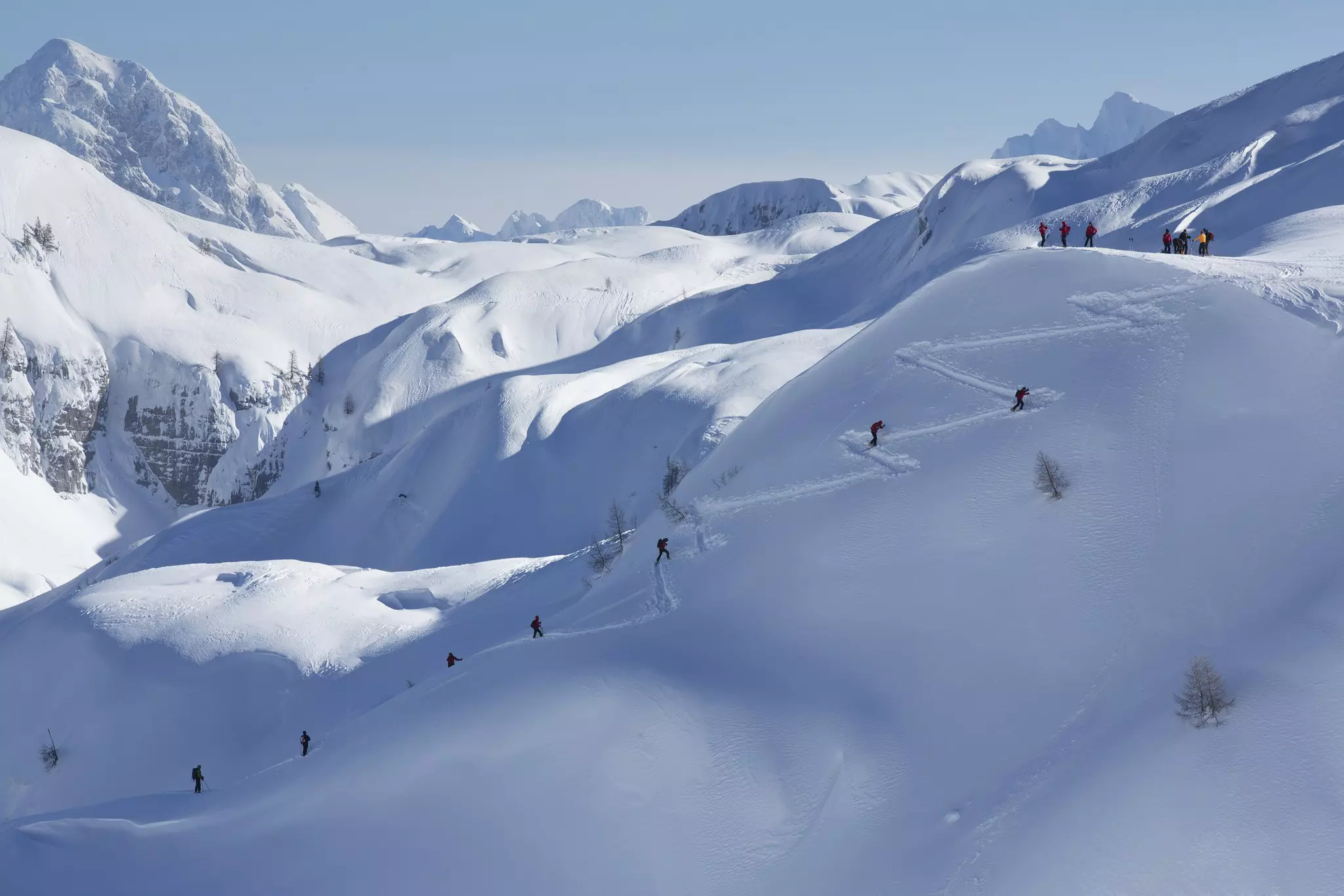 People hiking and snowshoeing up a single diagonal track in a snow-covered landscape of rounded hills, with mountain peaks in the background.