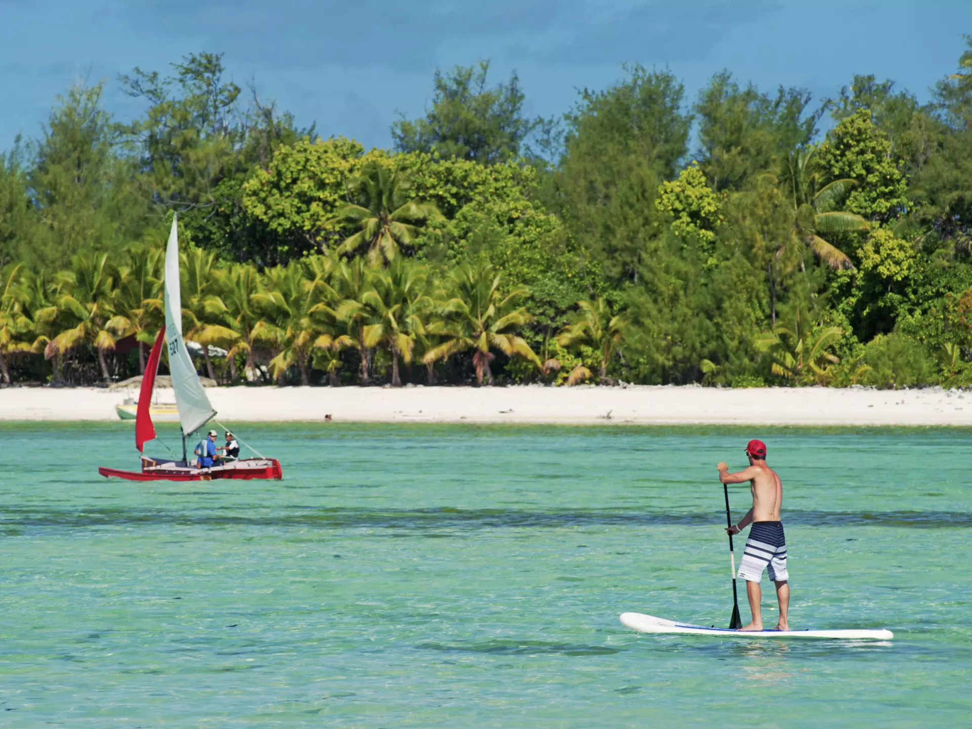 A man stand-up paddle-boarding on the crystal clear water of Muri Lagoon, with a small boat in the background