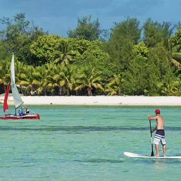 A man stand-up paddle-boarding on the crystal clear water of Muri Lagoon, with a small boat in the background