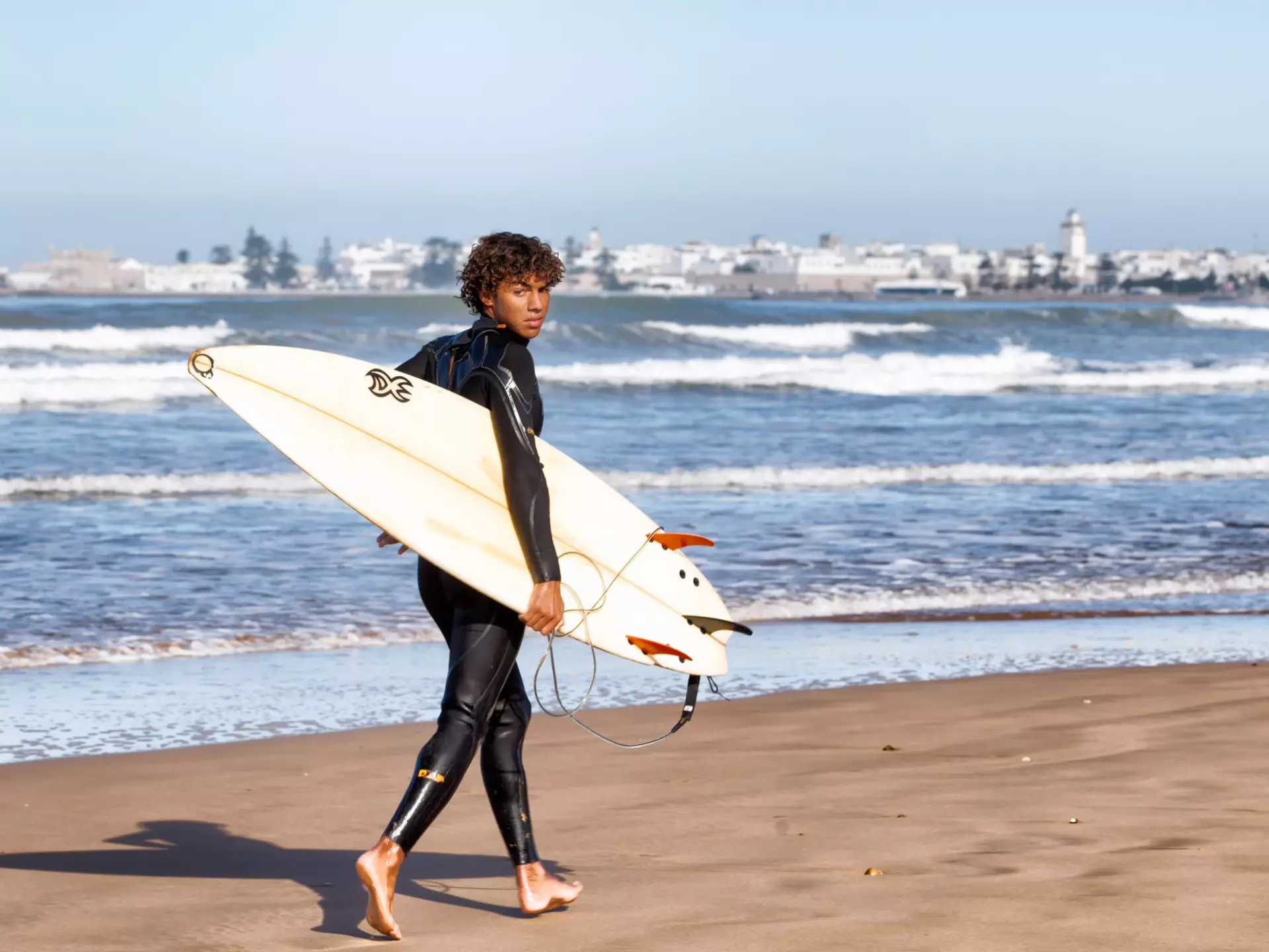 A surfer carrying his board looks at the camera as he walks along the beach.
