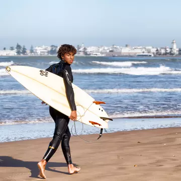 A surfer carrying his board looks at the camera as he walks along the beach.