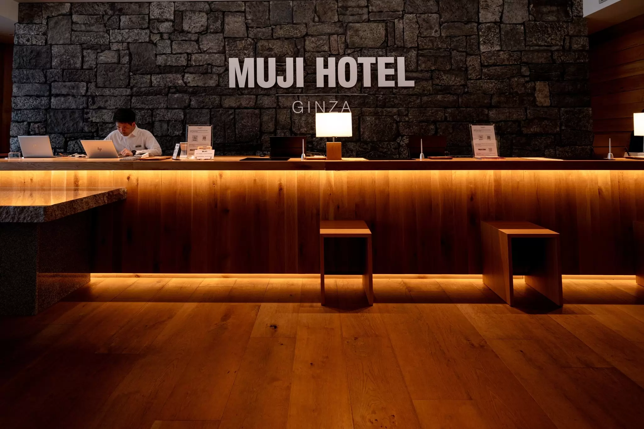 The large, wooden reception desk at a hotel with its name, "The Muji Hotel," in block letters on the wall behind it