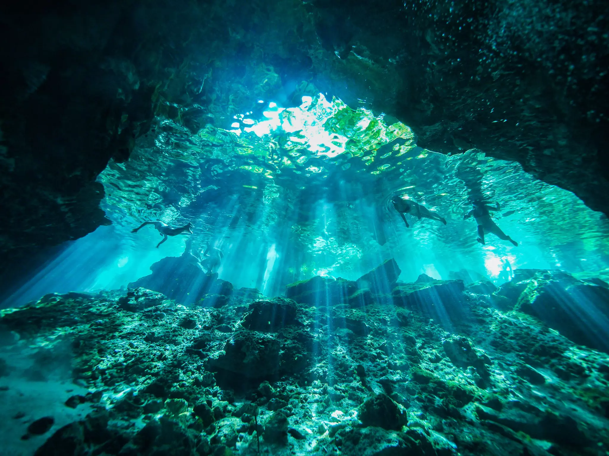 Underwater shot of three people swimming in an underwater cave with streaks of sunlight shining through.