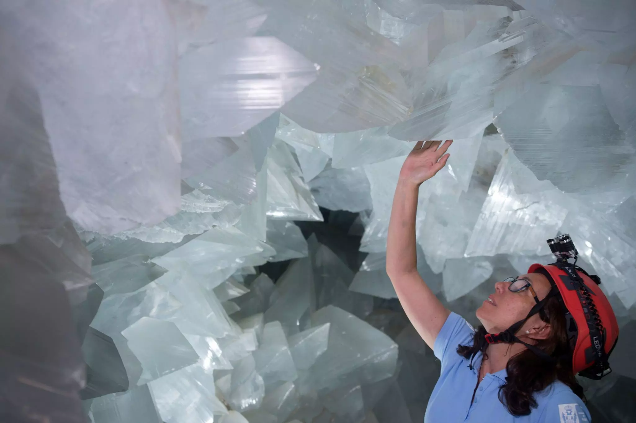 A woman wearing a red helmet with a torchlight attached touches a large white crystal descending from a ceiling. She's surrounded by large crystals inside the la geoda de pulpi