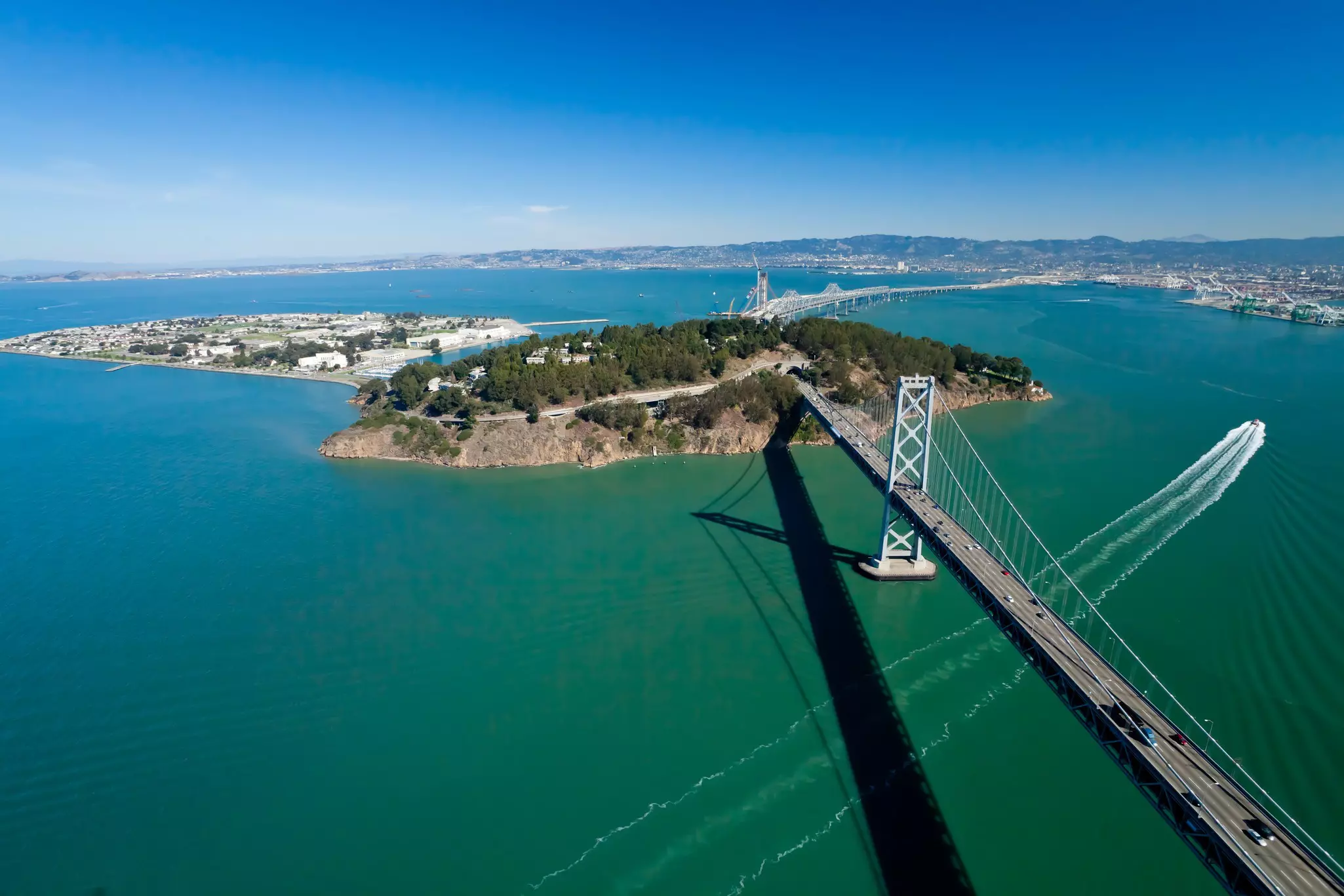 Aerial view of bridge leading to a forested island and then continuing beyond, surrounded by a bay on a sunny day.