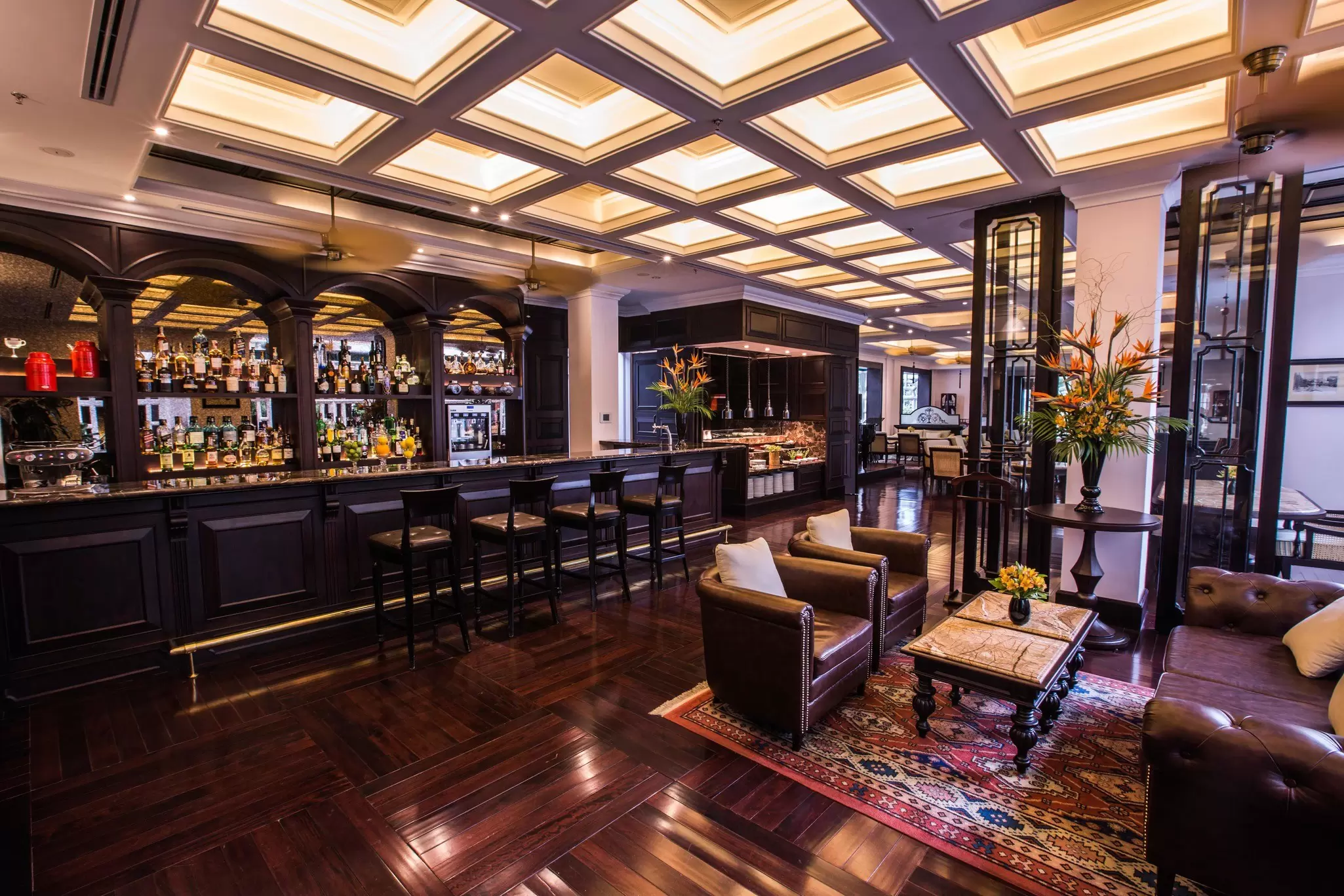 An elegant, bright lounge with dark wood floors, a long bar with high chairs. Small, soft individual chairs with pillows surround a coffee table with a small bouquet of flowers in the foreground