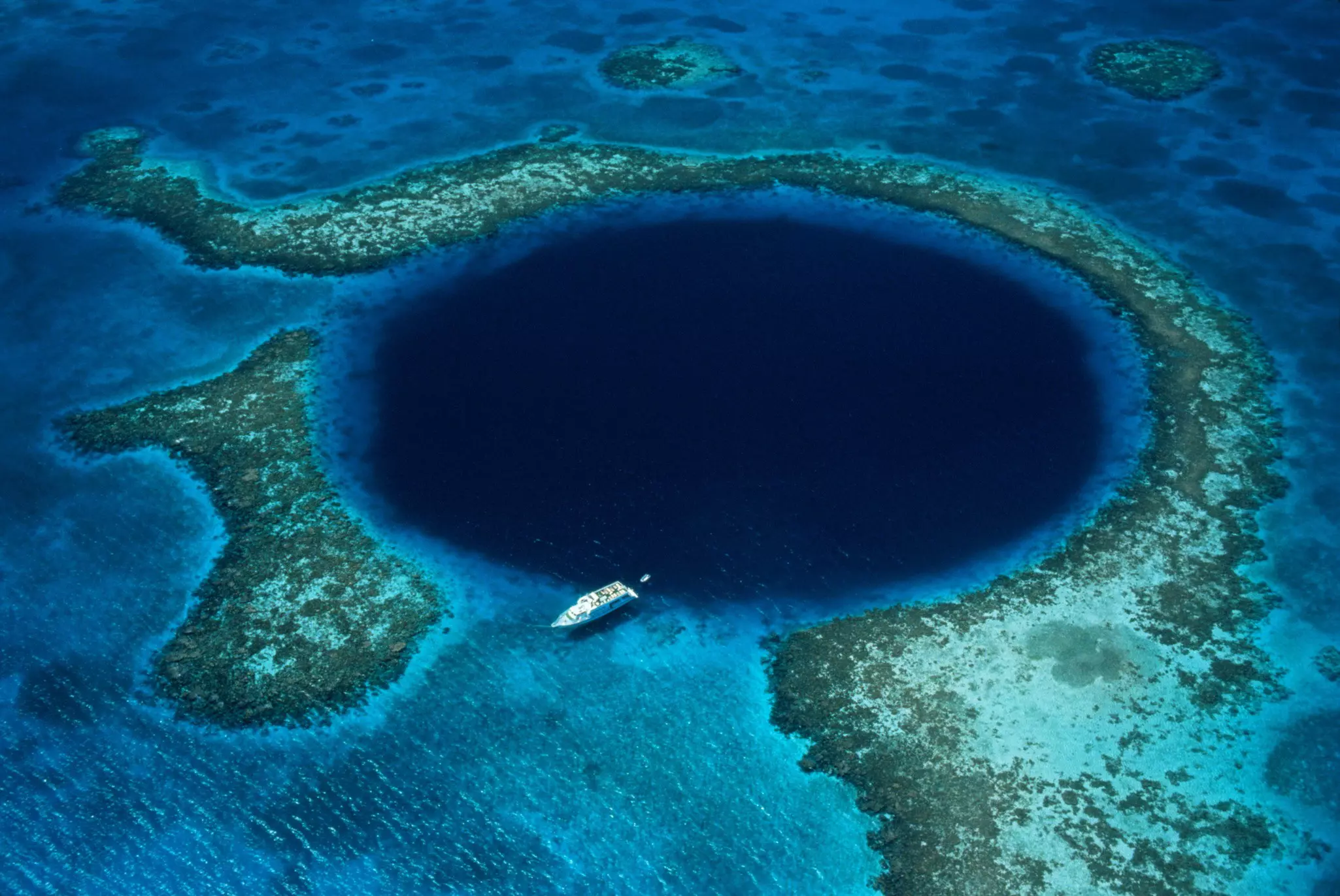 An aerial view of a boat next to the blue waters in a perfectly round sinkhole surrounded by lighter-colored reefs and sand.
