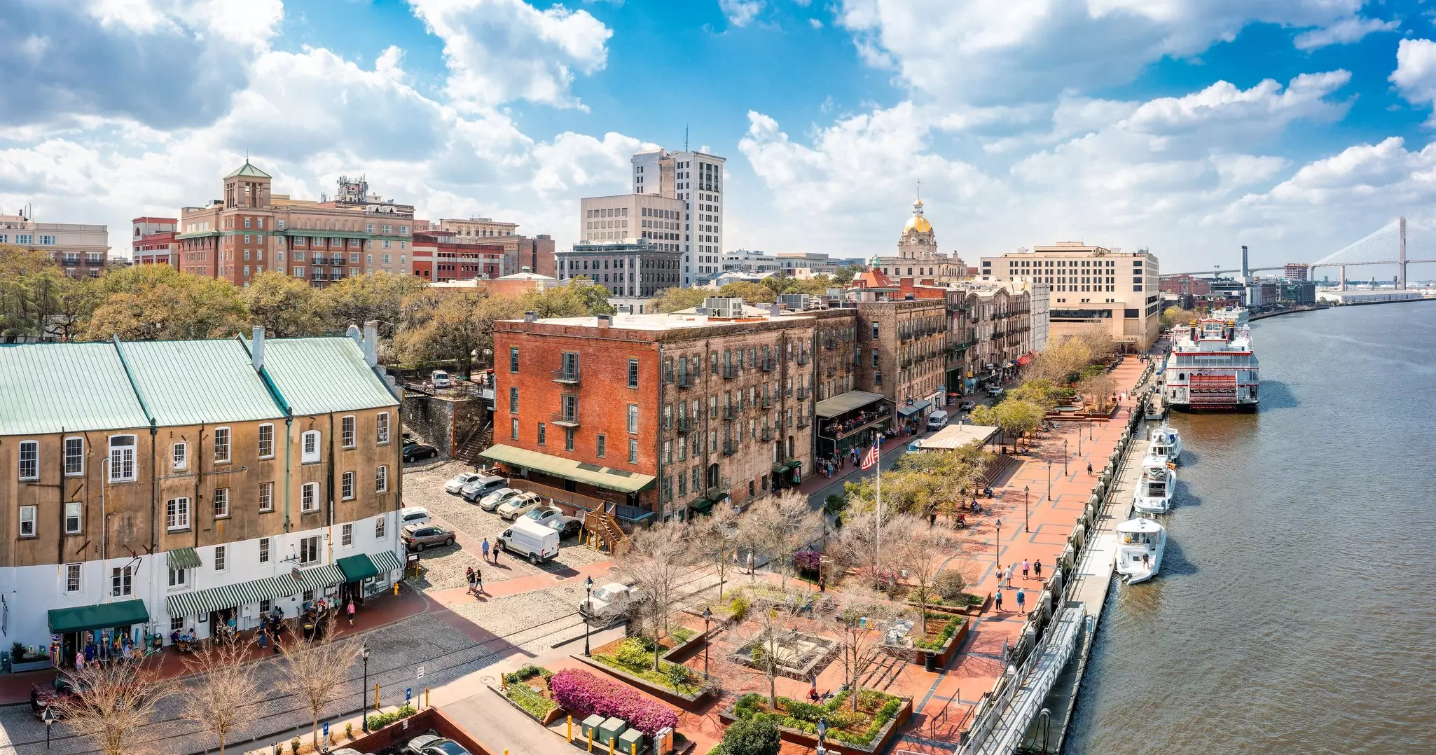 An aerial view of Savannah, Georgia, along the river; a few tall buildings and a building with a gold dome are visible in the distance, as is a suspension bridge.