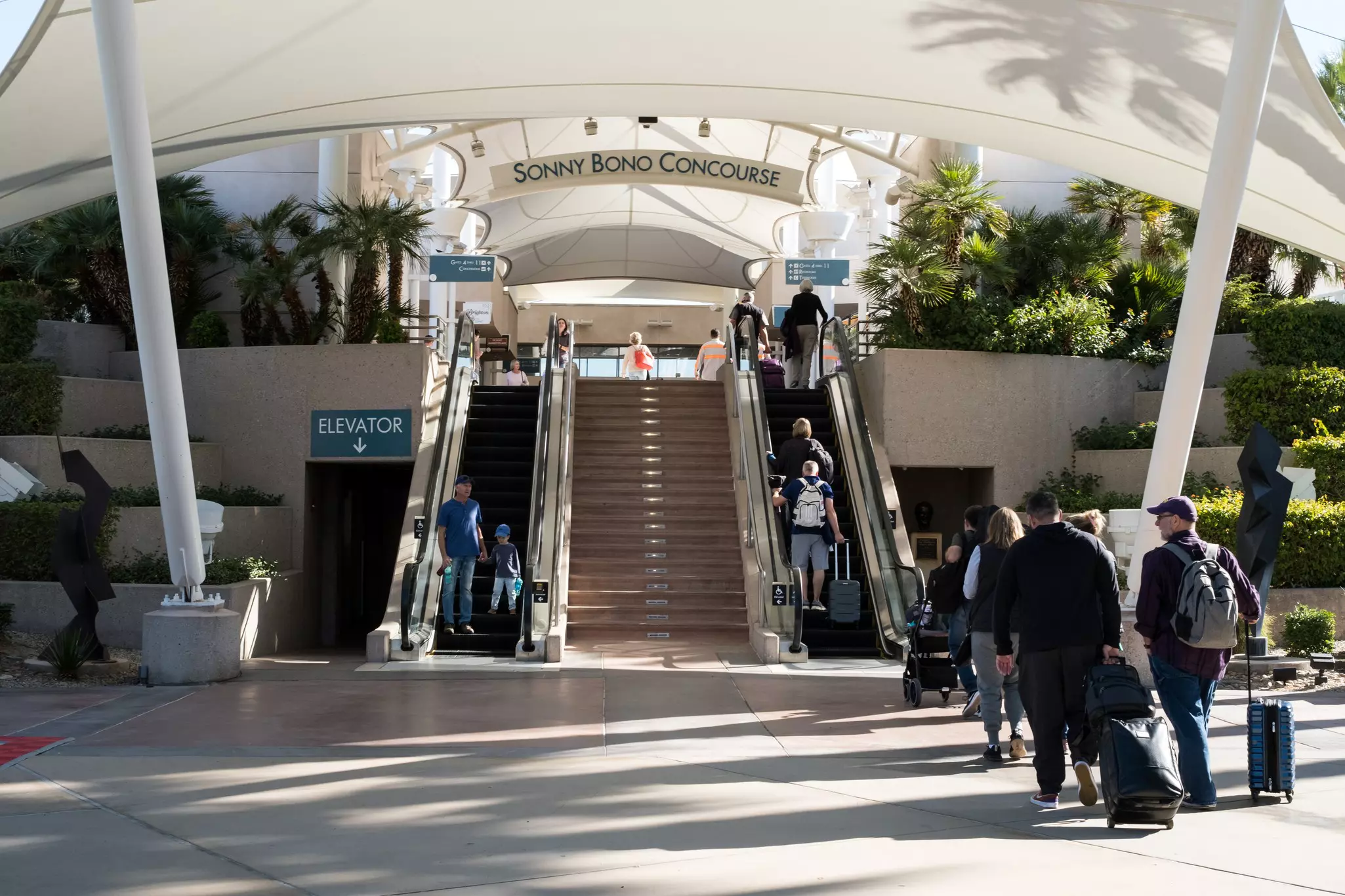 Travelers use two escalators up to an airport concourse, with an overhead sign reading "Sonny Bond Concourse," framed by palm trees