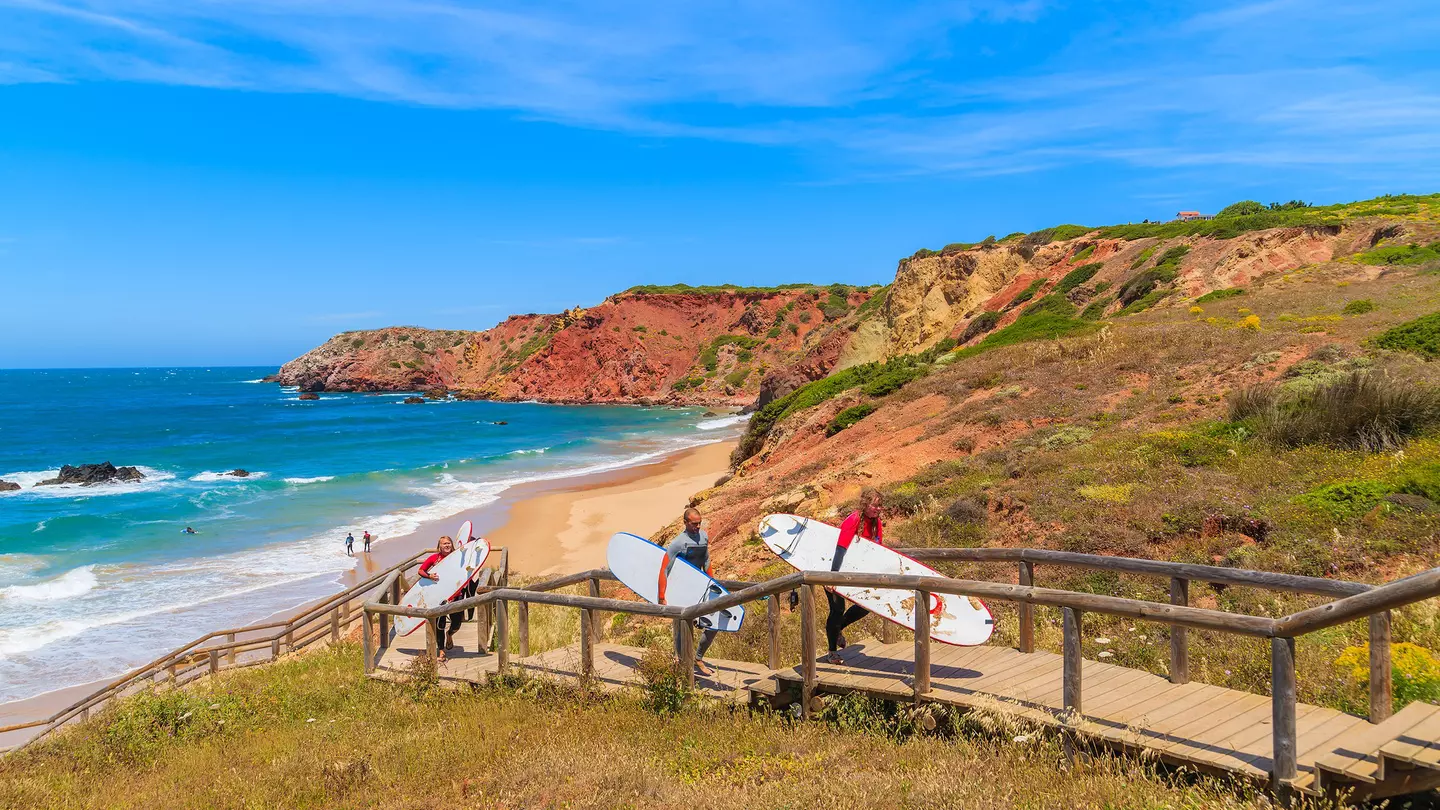 Surfers holding surfboards walk on a footbridge from a rocky, cliff-lined beach in the Algarve region