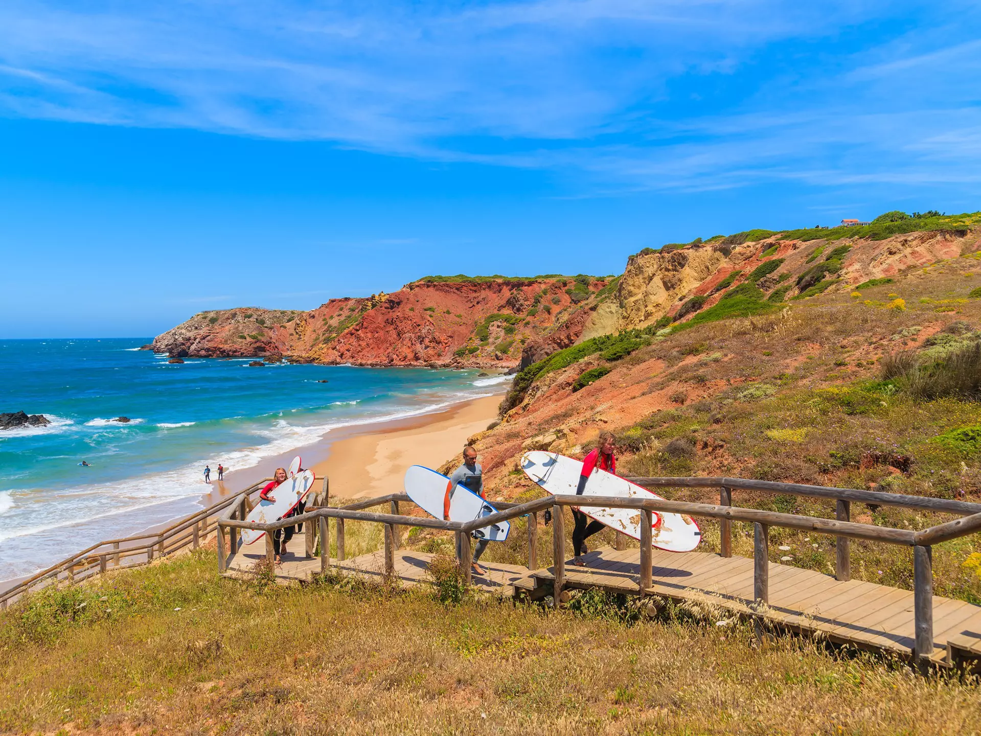 Surfers holding surfboards walk on a footbridge from a rocky, cliff-lined beach in the Algarve region