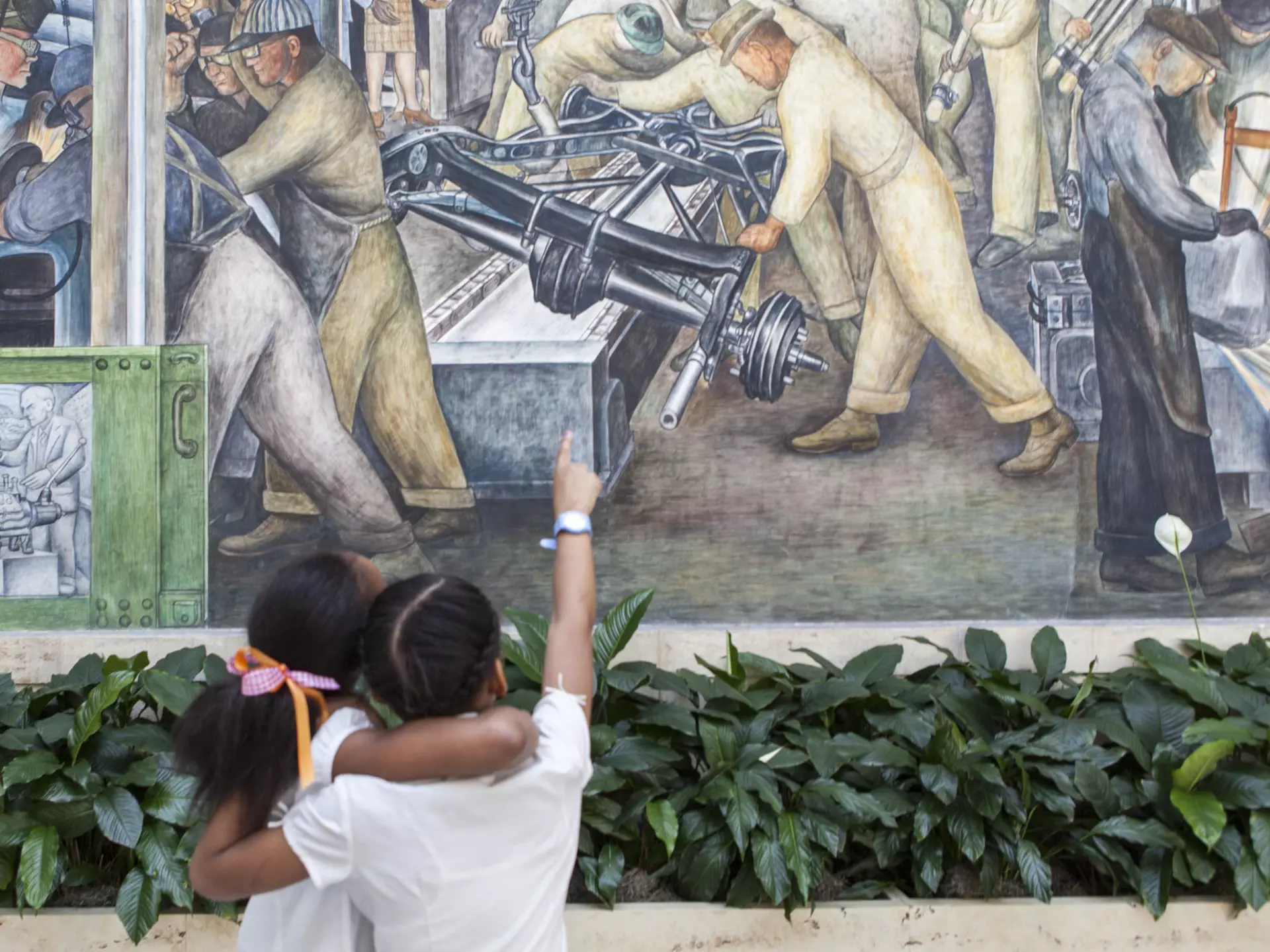 Two young children with arms around each other and one pointing up at a large mural by Diego Rivera with green peace lily flowers in front inside a museum.