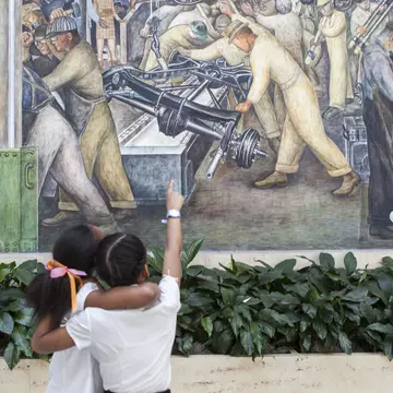 Two young children with arms around each other and one pointing up at a large mural by Diego Rivera with green peace lily flowers in front inside a museum.