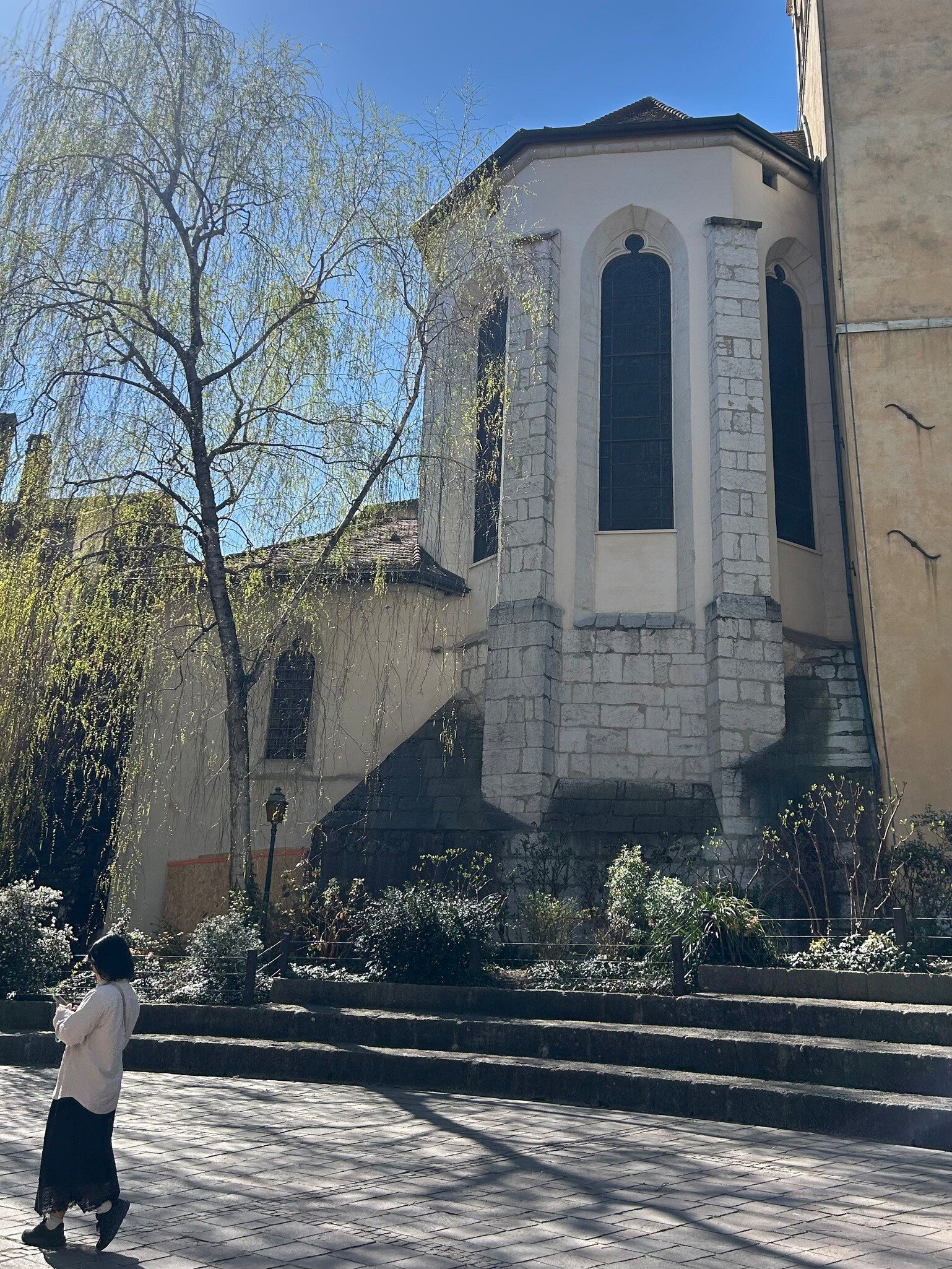 Stone apse of a Gothic church with tall windows between buttresses, a weeping willow and a passer-by below.
