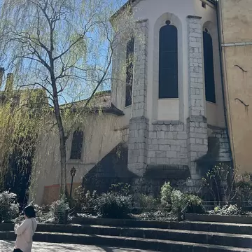 Stone apse of a Gothic church with tall windows between buttresses, a weeping willow and a passer-by below.