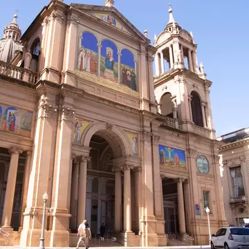 A tall sandstone cathedral with colourful religious mosaics above its arches, a pedestrian passing on the street below.