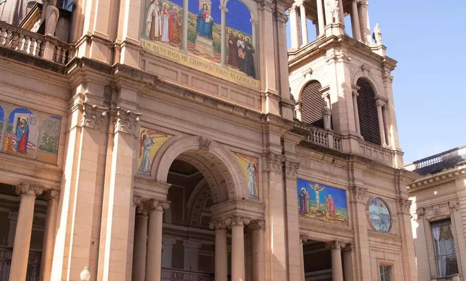 A tall sandstone cathedral with colourful religious mosaics above its arches, a pedestrian passing on the street below.