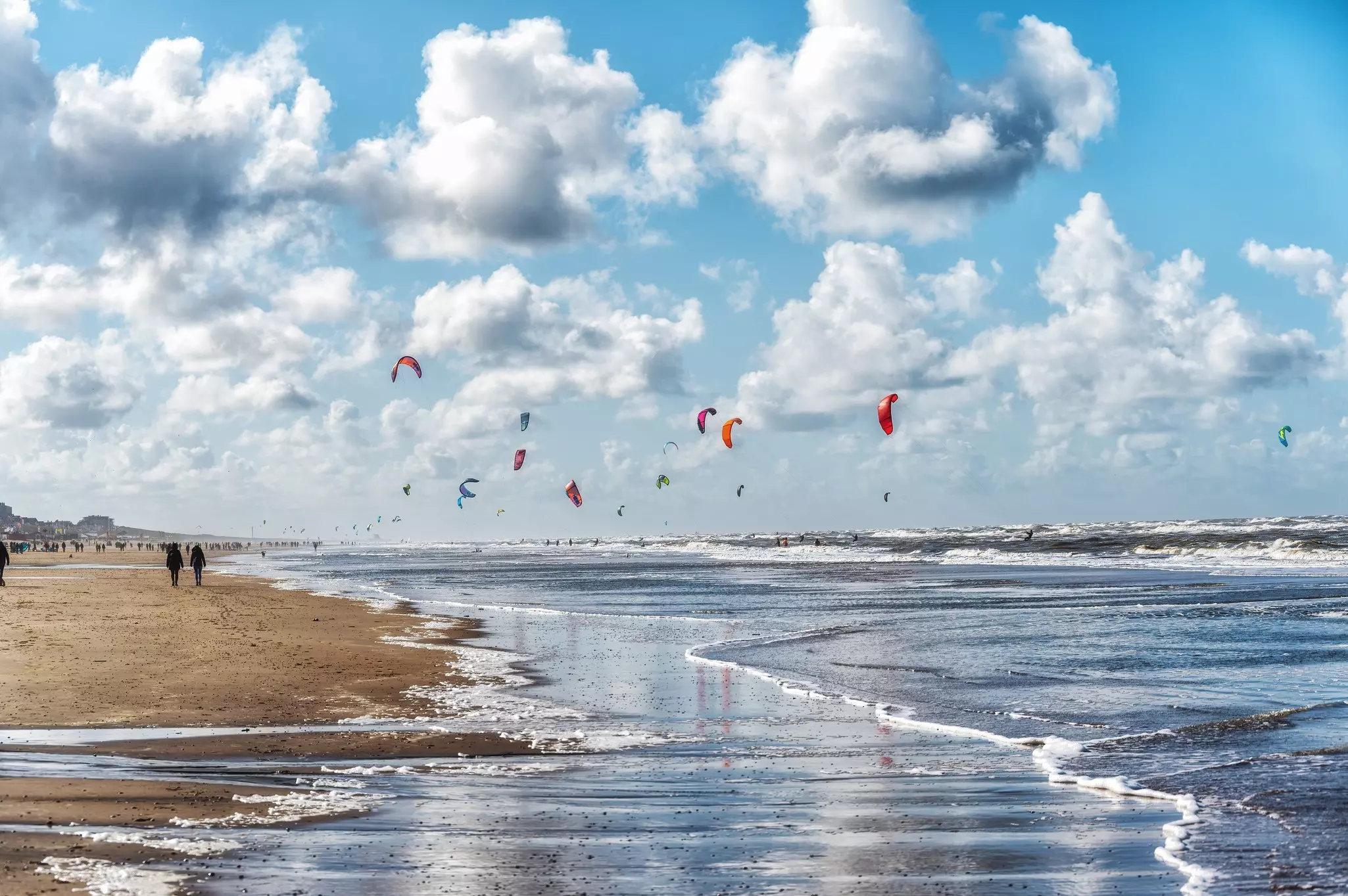 Waves lap at the shores of a beach busy with walkers on a cool day. Kites fill the sky above the kitesurfers out on the water.