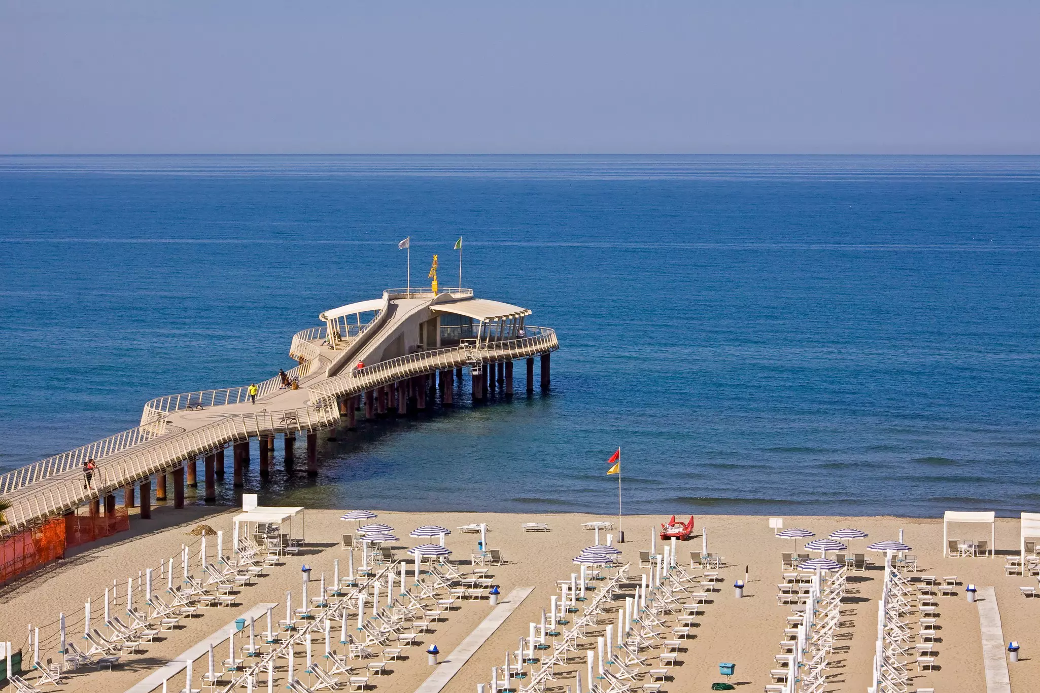 A beach lined with empty chairs and umbrellas on a sunny day with a pier stretching into the ocean.
