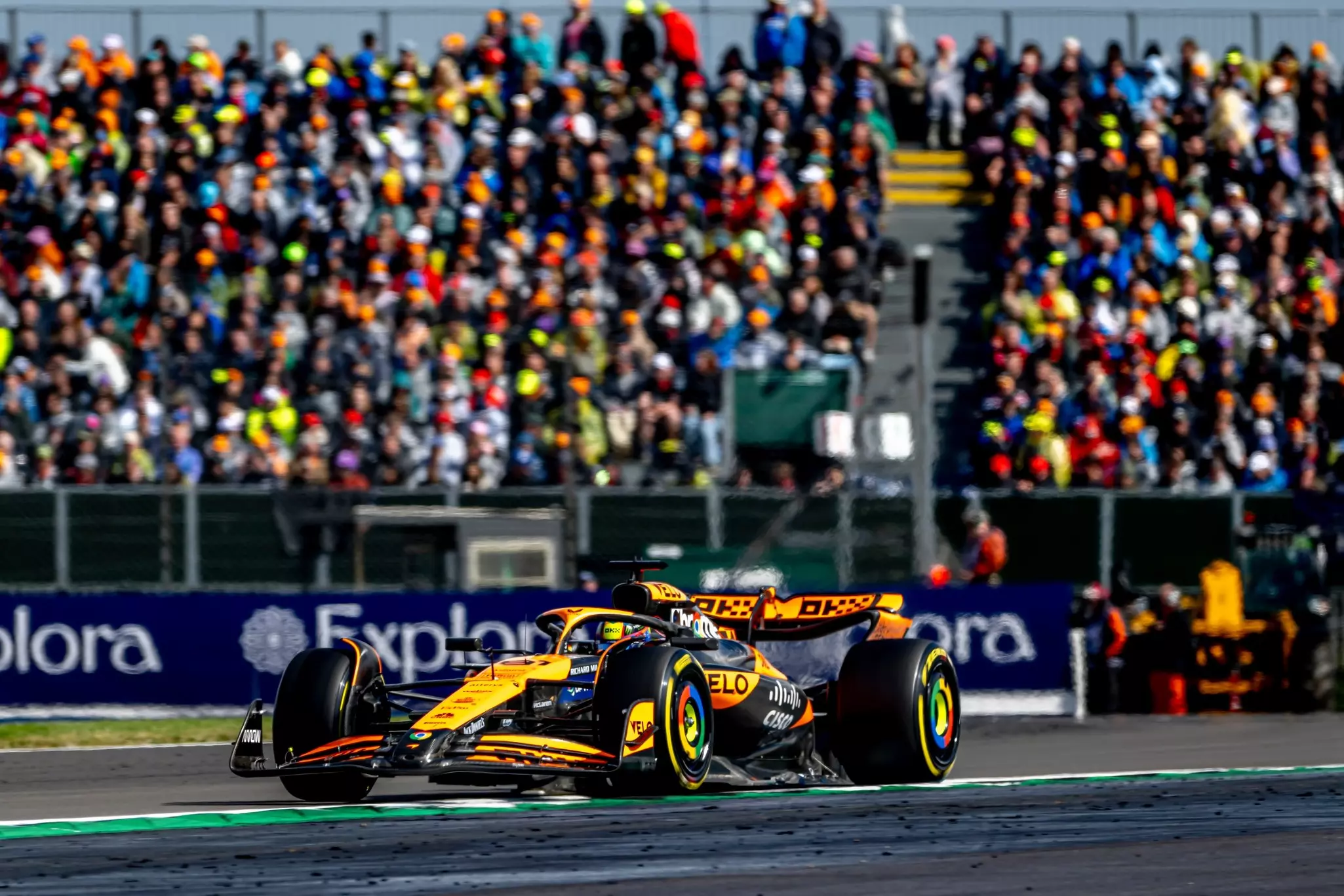 A yellow racing car drives on a track, with thousands of spectators watching from bleachers.