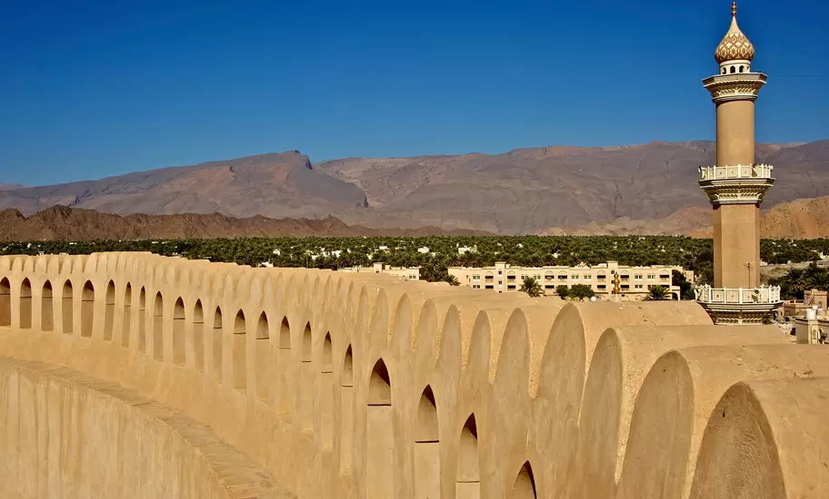 Arched sandstone parapet of Nizwa Fort's round tower, a mosque minaret, palm groves and the Hajar Mountains behind