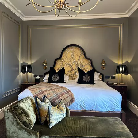 A bed with a white covering has a plaid blanket laid on top and decorative black pillows. The triangular headboard is golden fabric, the bedside tables have lamps, and the walls are gray.