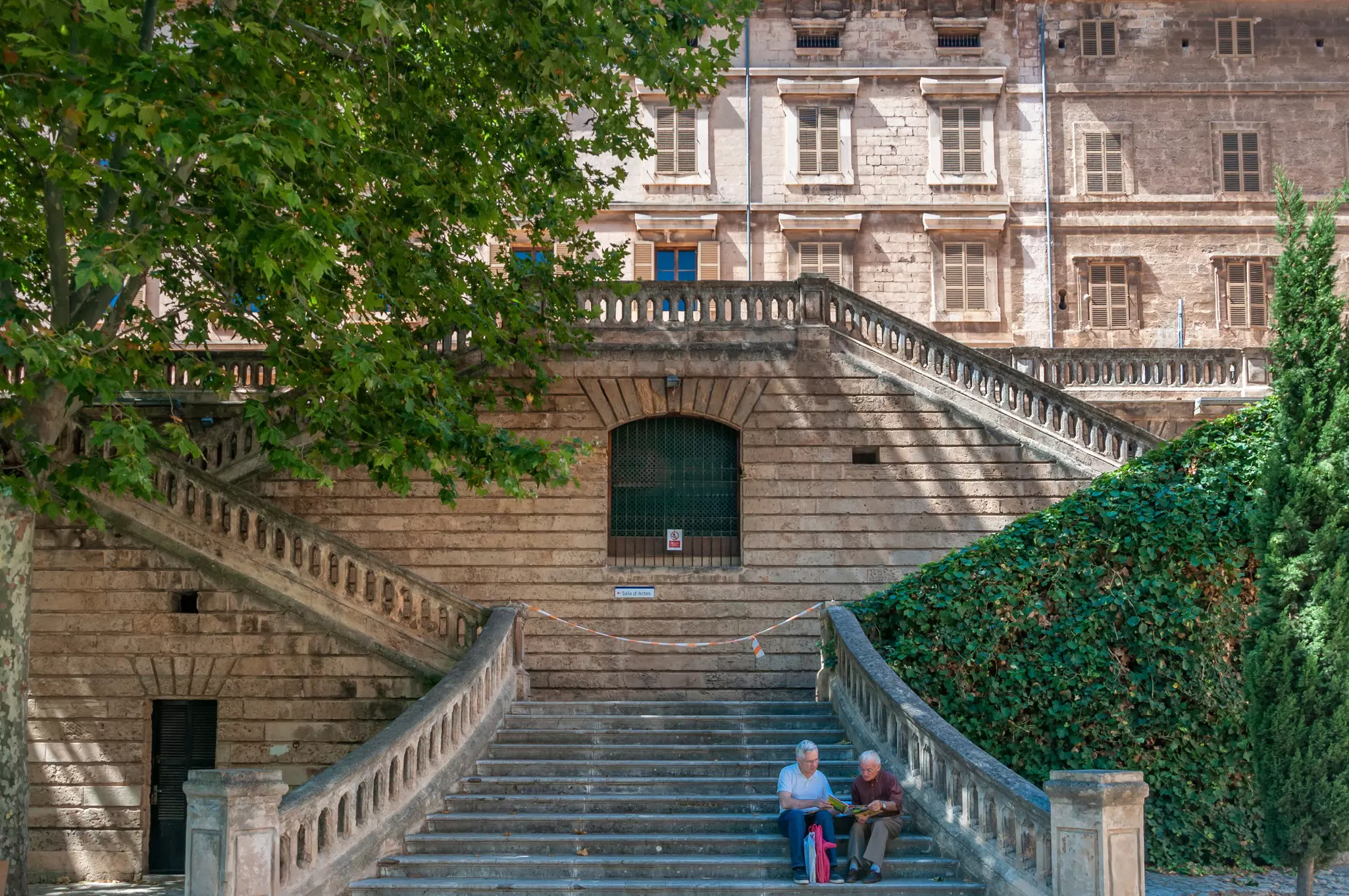 Two people sit on a stone staircase, shaded by trees, on a city street.