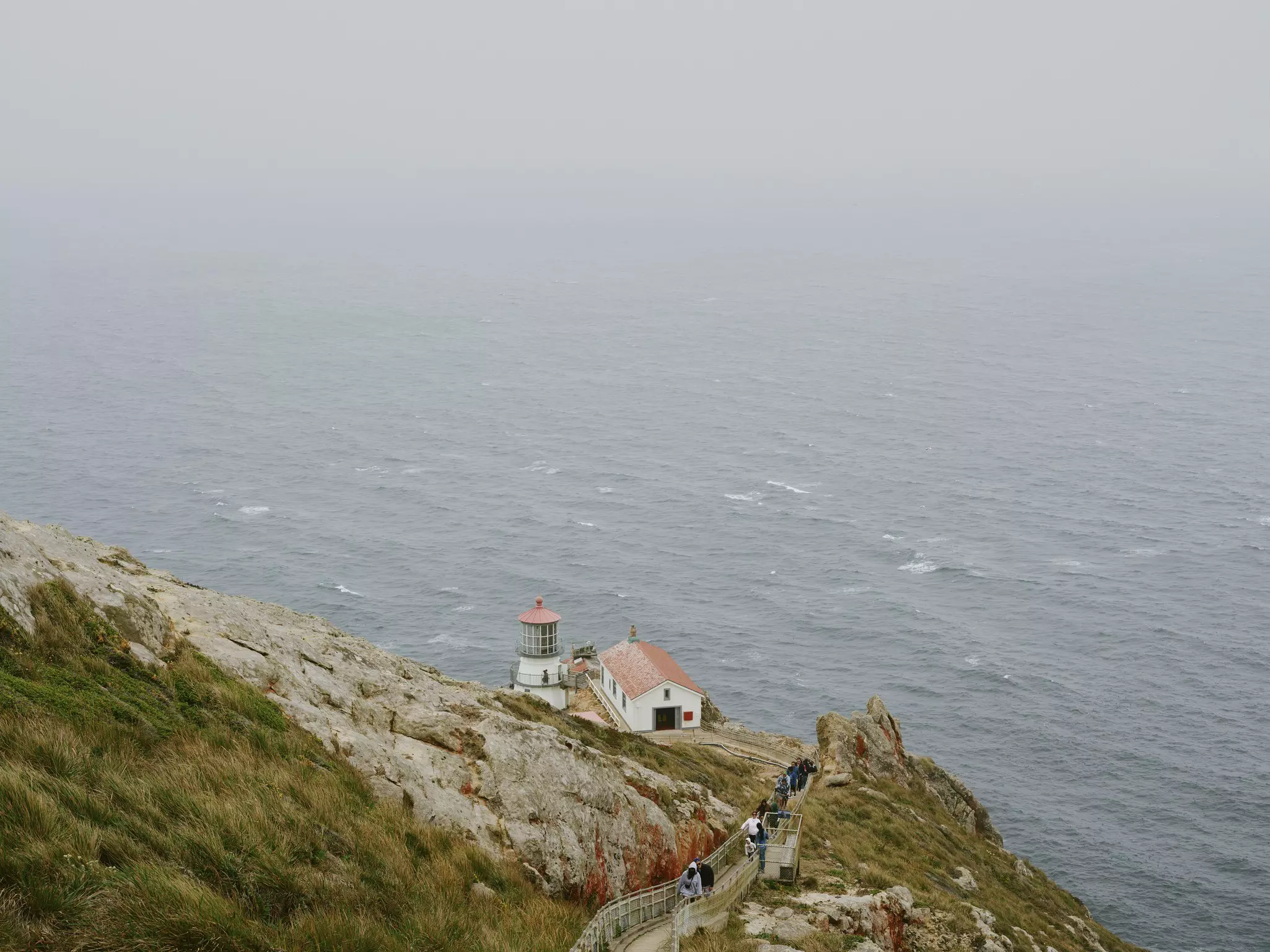 Steps down to a rocky cliff toward a red-roofed lighthouse, the sea in the background