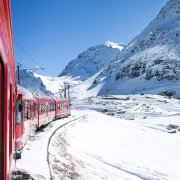 Red passenger train curving along a snowy track beneath steep white mountains and a clear blue sky in the Swiss Alps.