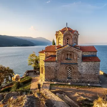 Brick-and-stone Byzantine church with red-tiled domes on a clifftop terrace, lake and hazy hills beyond in late sun