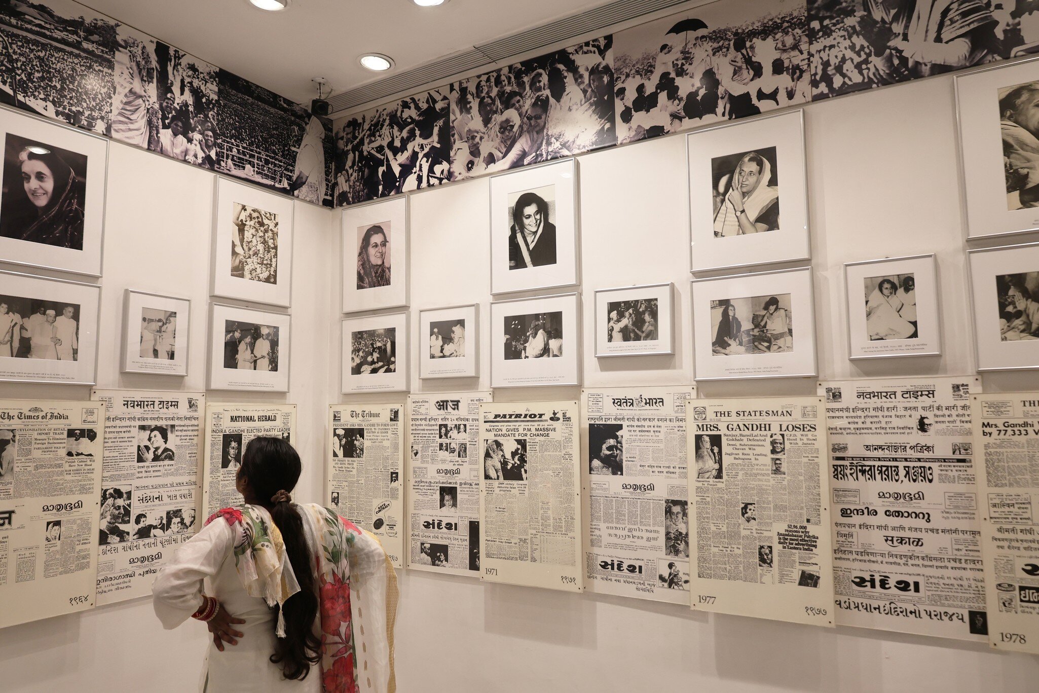 Woman viewing a wall display of framed historical photographs and newspaper clippings related to Indira Gandhi's political career. New Delhi, India.
