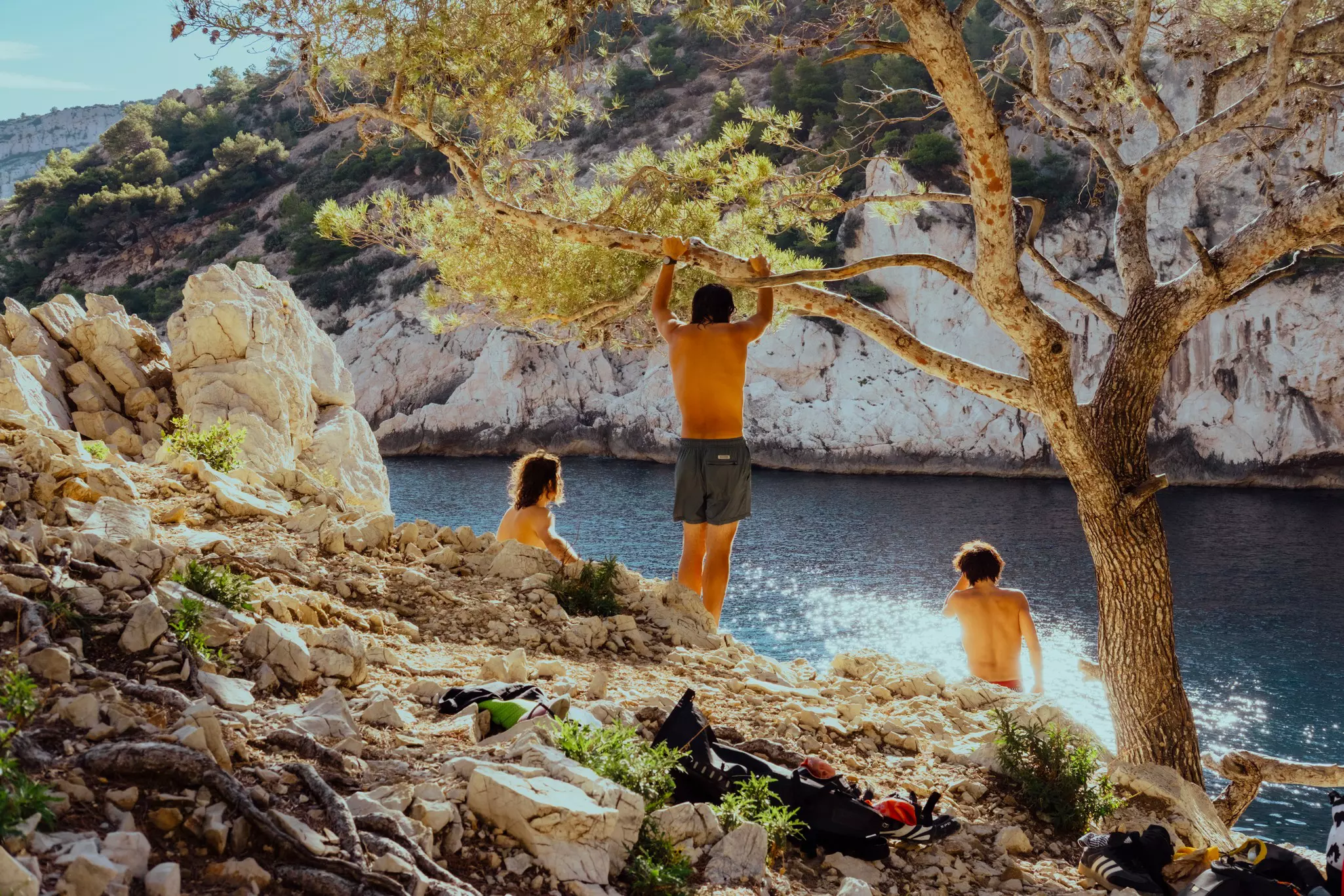 Three people on a rocky cliff, one holding on to a tree branch; light shines on the water.