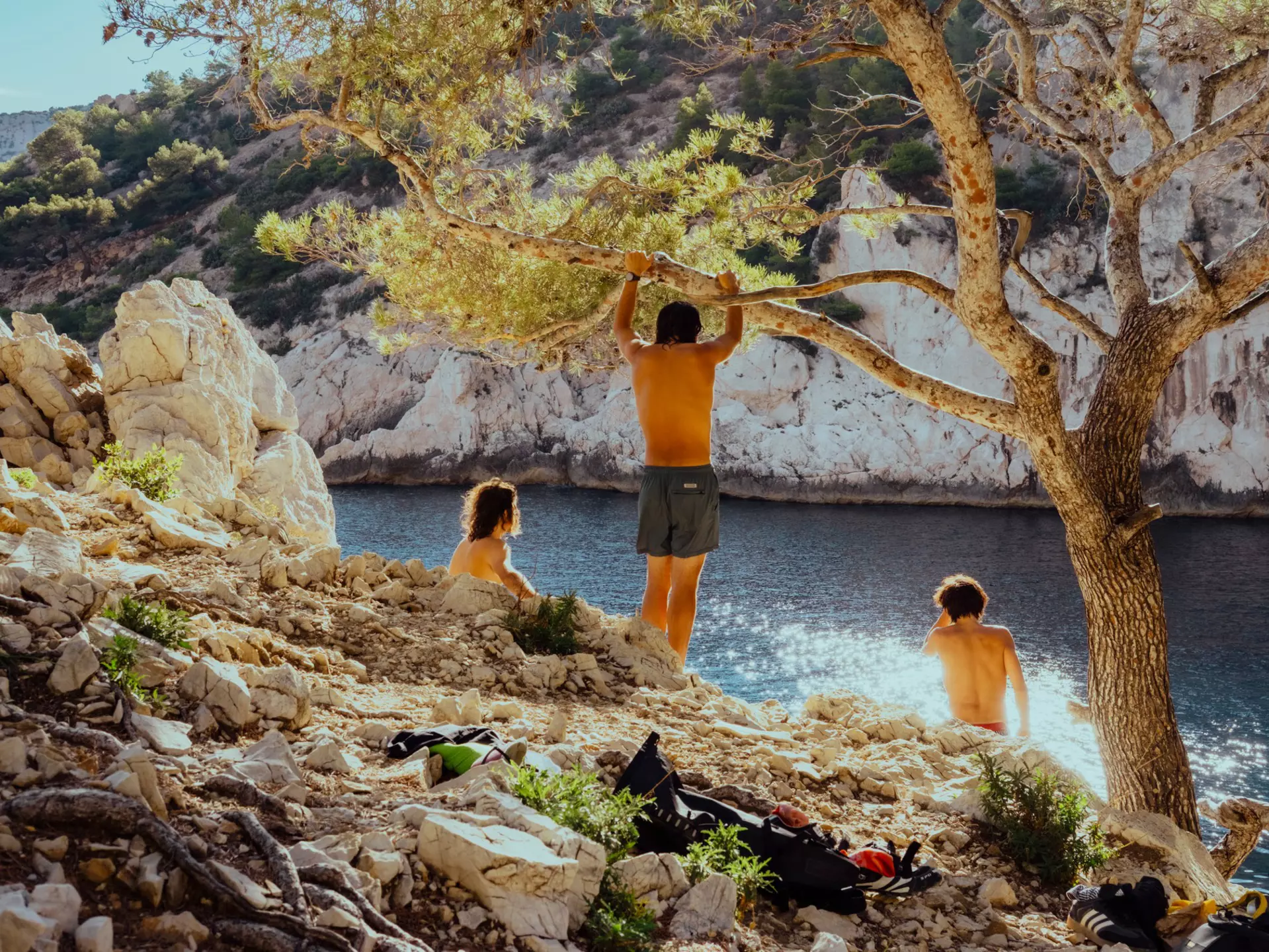 Three people relax in the shade of a tree near a cliff-backed beach on a sunny day