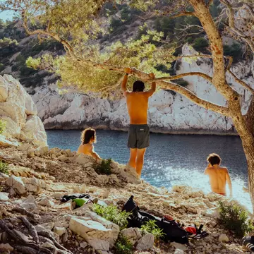 Three people relax in the shade of a tree near a cliff-backed beach on a sunny day