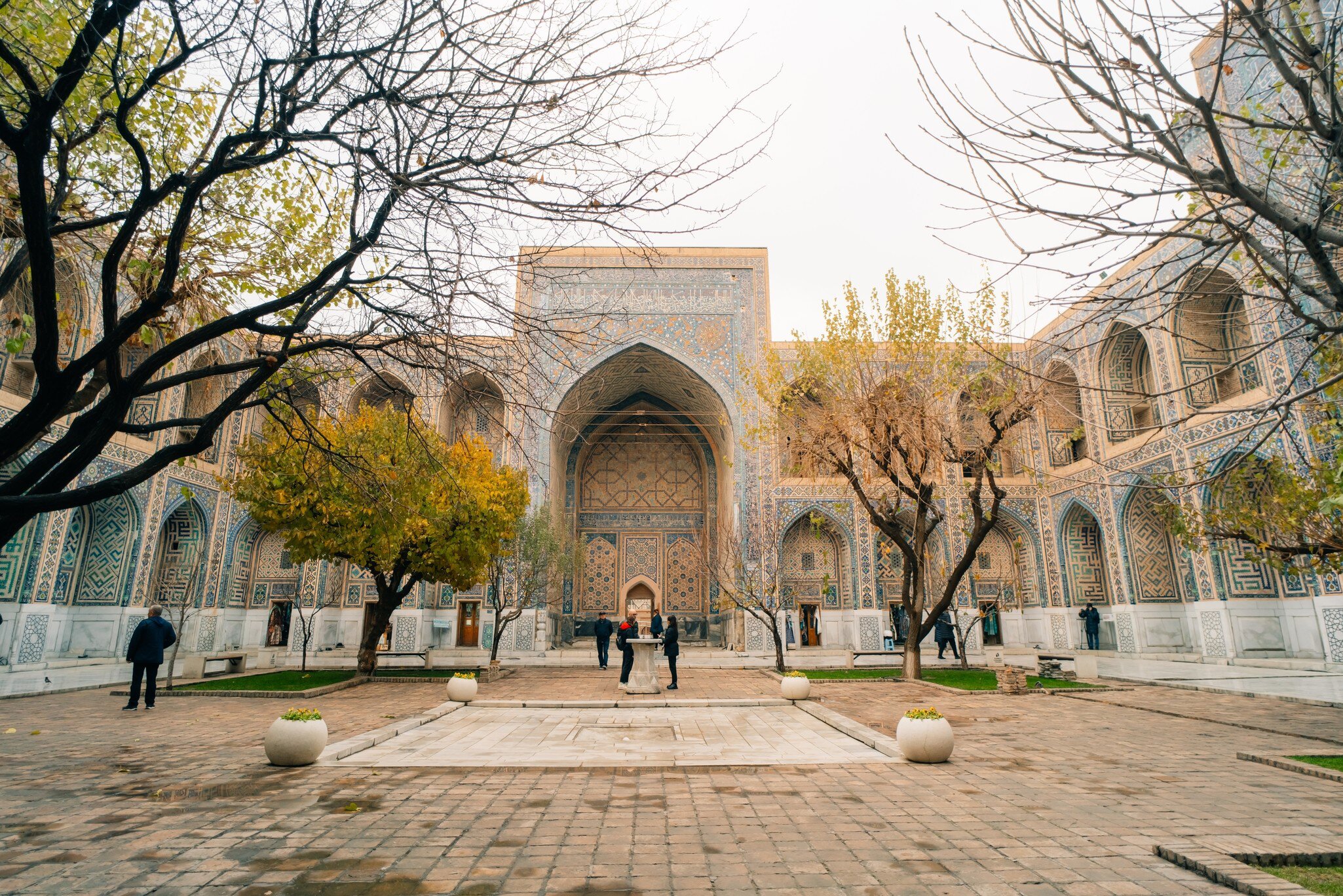 Tiled courtyard of a madrasah on Registan Square, Samarkand, Uzbekistan, with autumn trees and a few visitors
