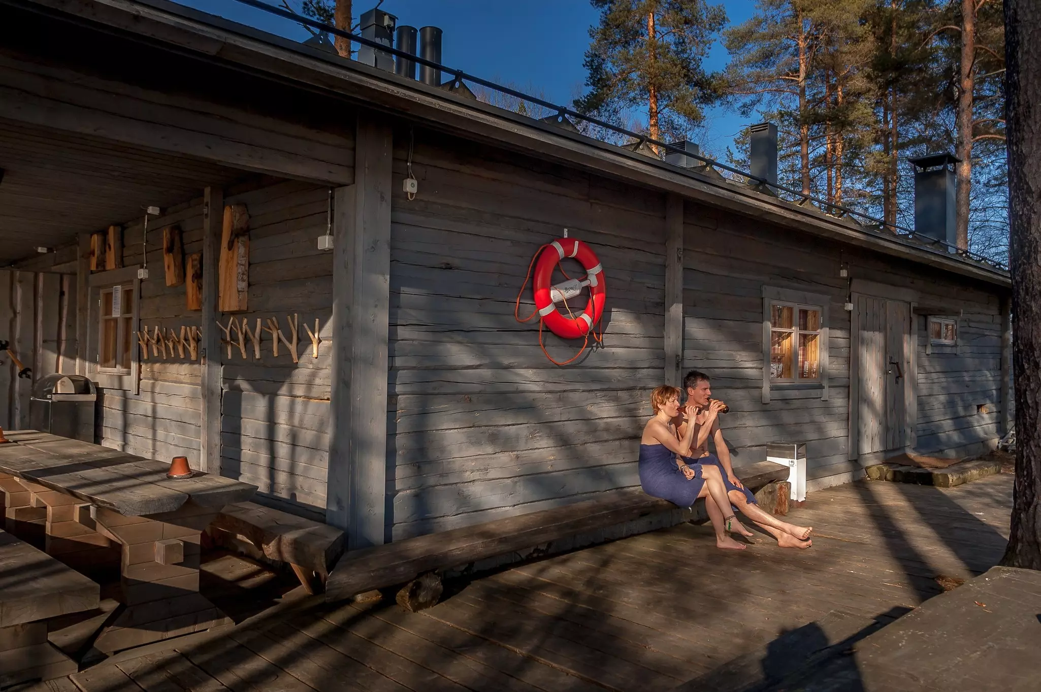Two people sit on a wooden bench leaning agains a wooden building with an orange ring hanging from it drinking bottles of beer while wrapped in towels.