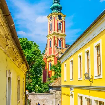 Walkers on a narrow cobbled lane between yellow houses, a red and yellow church tower with a green spire ahead, Szentendre.