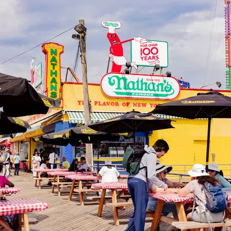 People at picnic tables with red-and-white checked tablecloths in front of a sign that reads "Nathans'" in green.