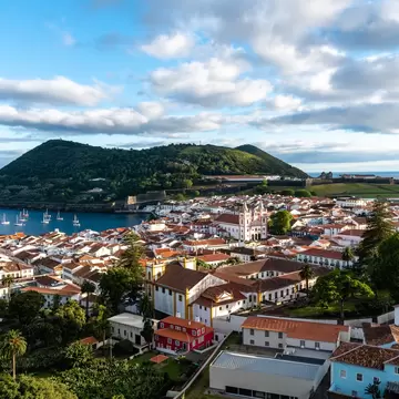 Aerial view of town of mostly white buildings with reddish roofs, with a bay and forested hillside in the distance on a sunny day.