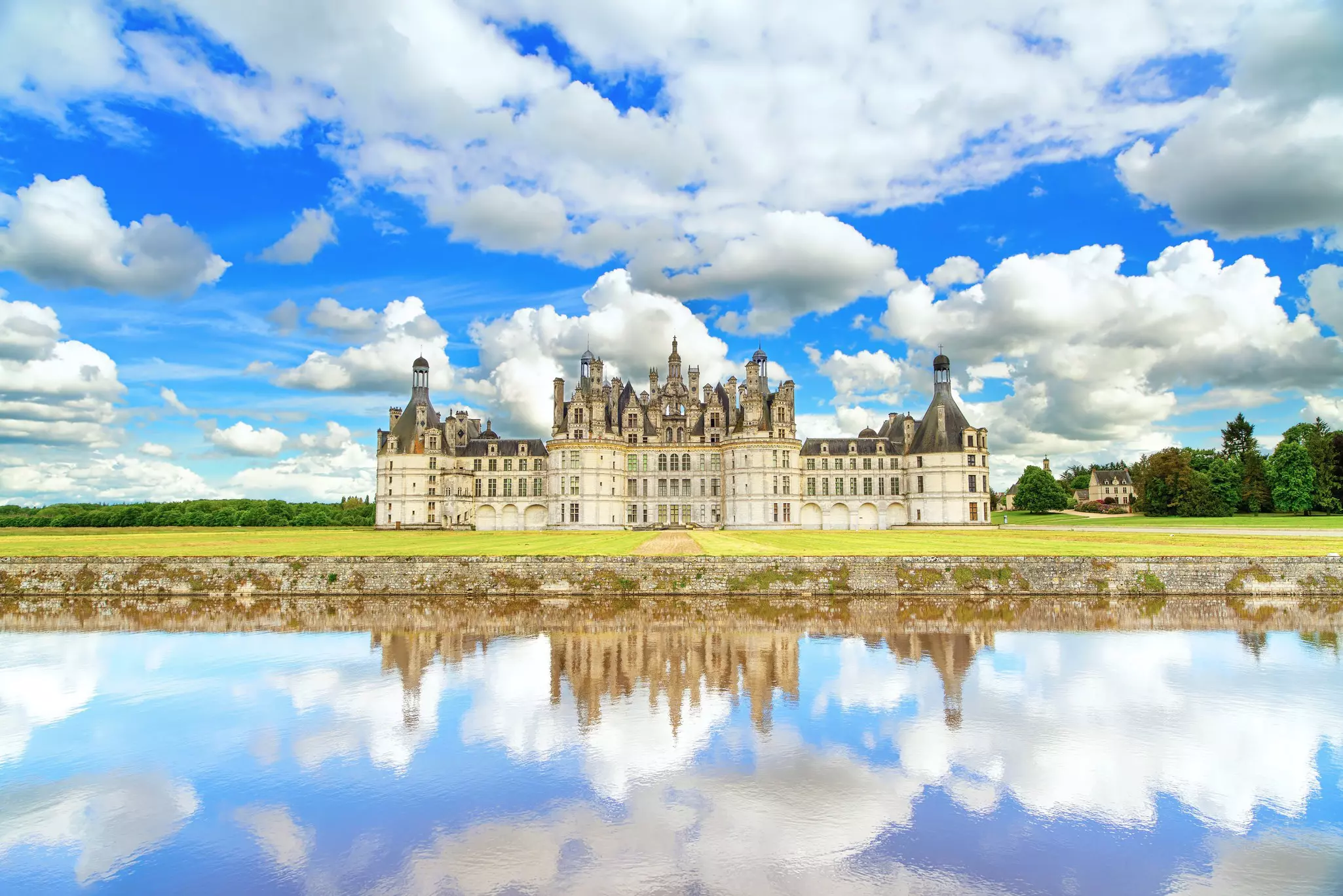 A vast white-brick renaissance building with turrets and a grey slate roof reflected in a large body of still water that perfectly mirrors the cloudy blue sky above