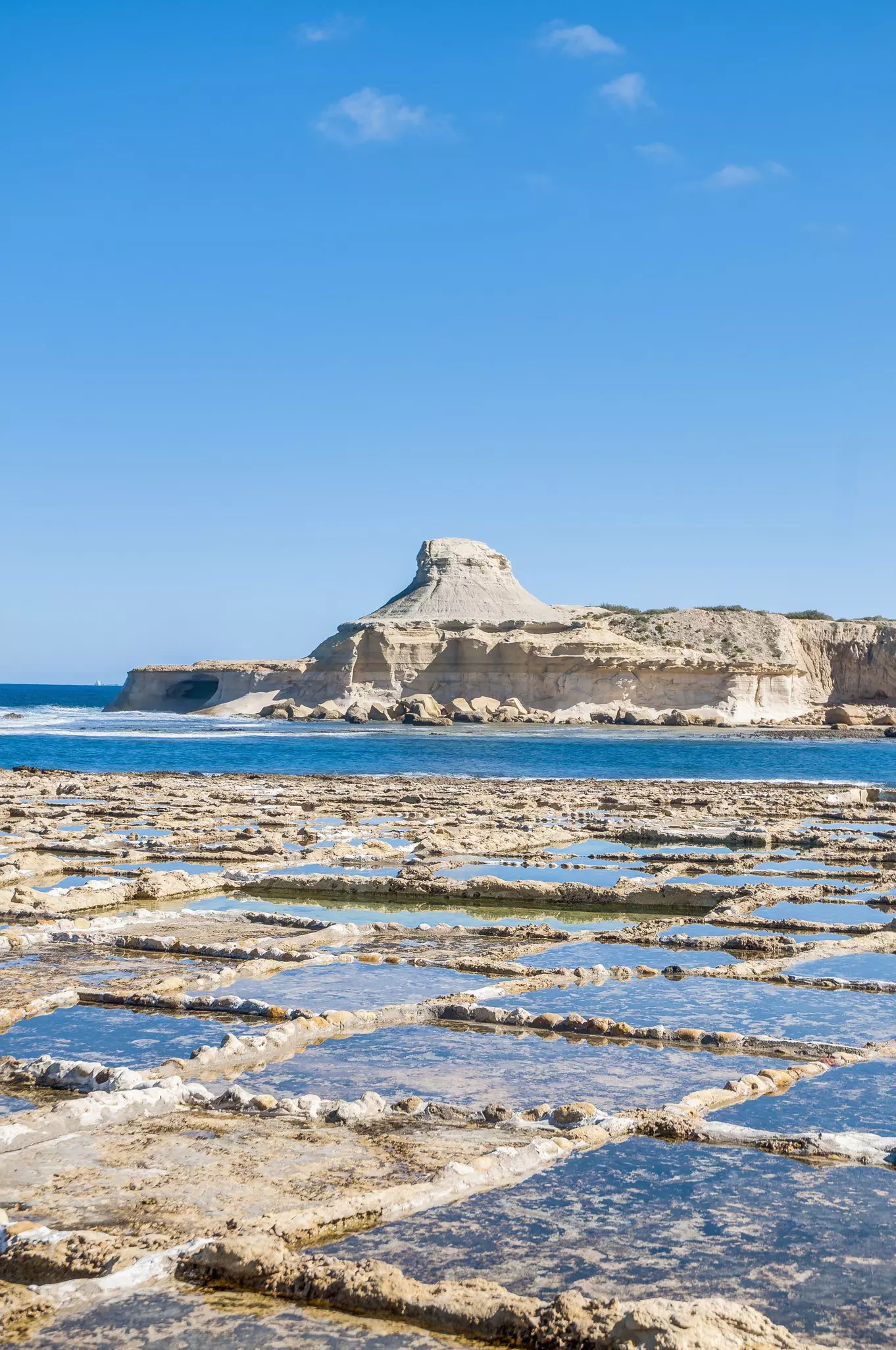Small, shallow rectangular ponds hewn into the flat limestone bed are filled with water. The sea lies beyond, as does an pyramidal rock outcrop of bleached stone