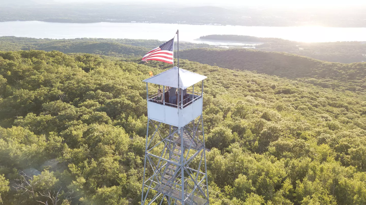 A woman in a tower with an American flag, looking out over the Hudson River, greenery and mountains