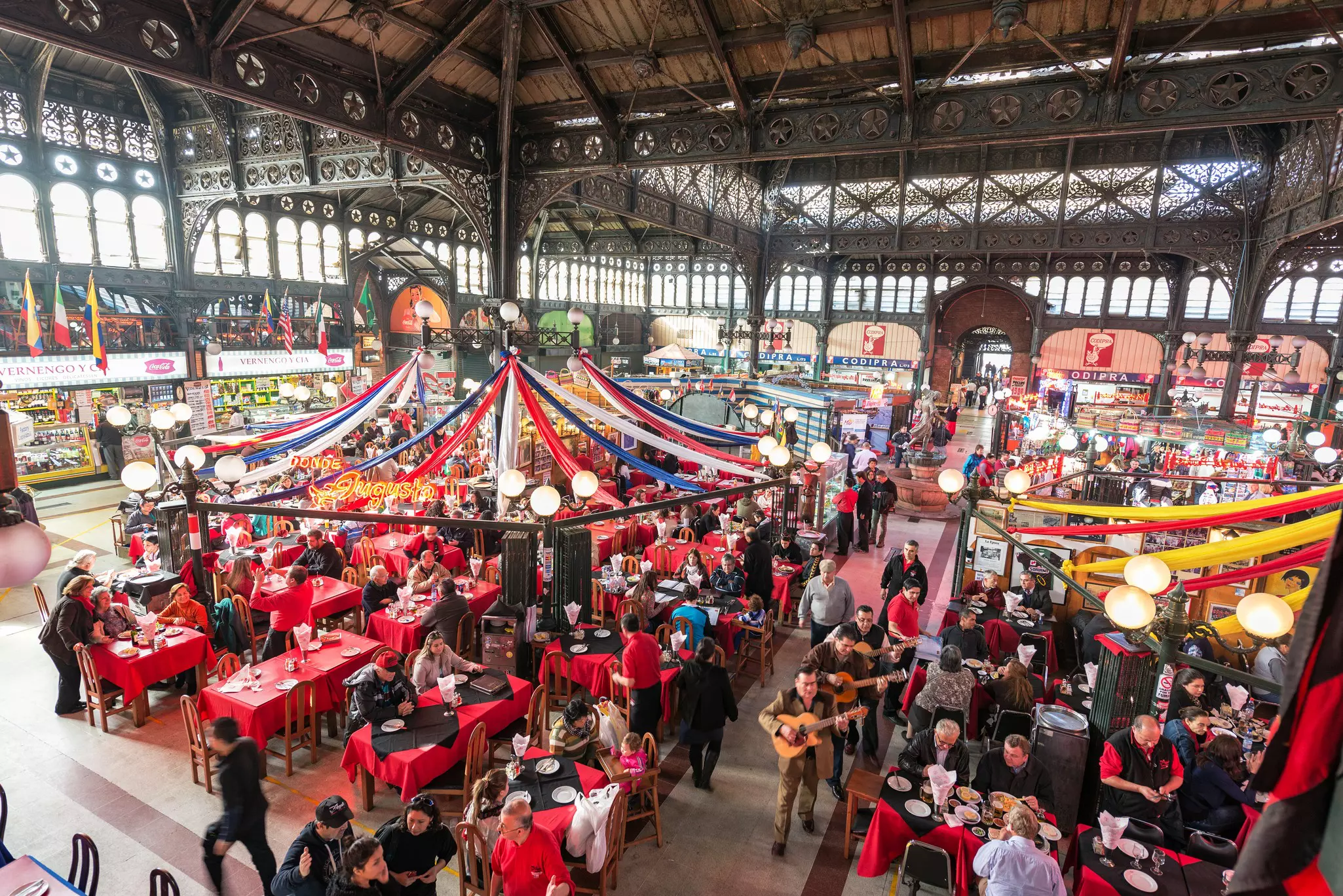 The view of an interior of a market, filled with tables and covered by wrought-iron beams.