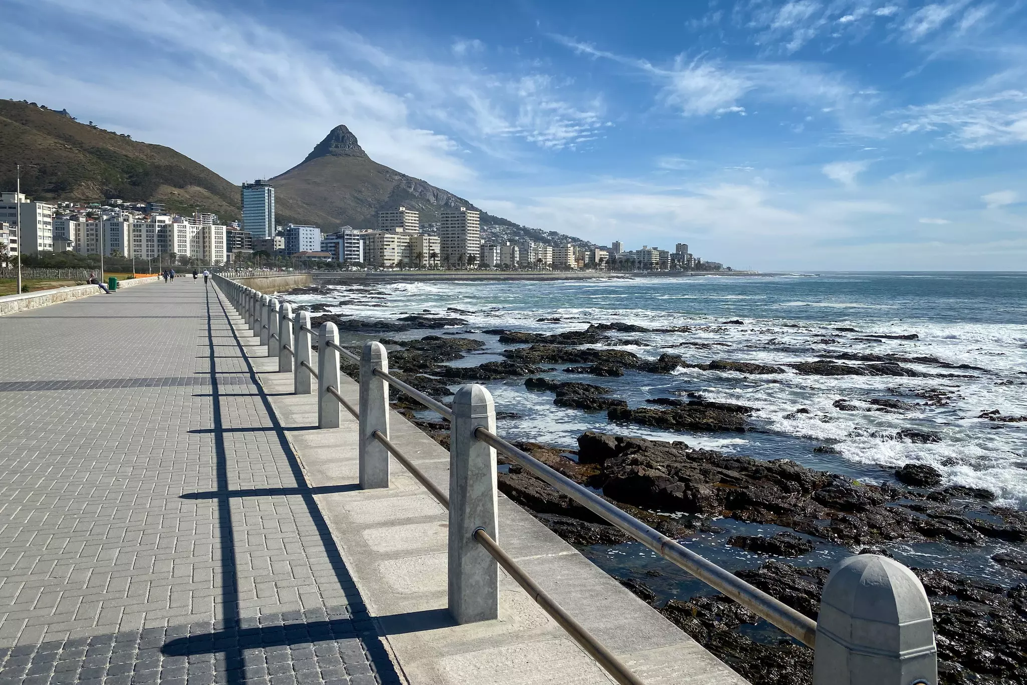 A paved walkway with a railing by the sea, with a mountain and tall buildings in the distance.