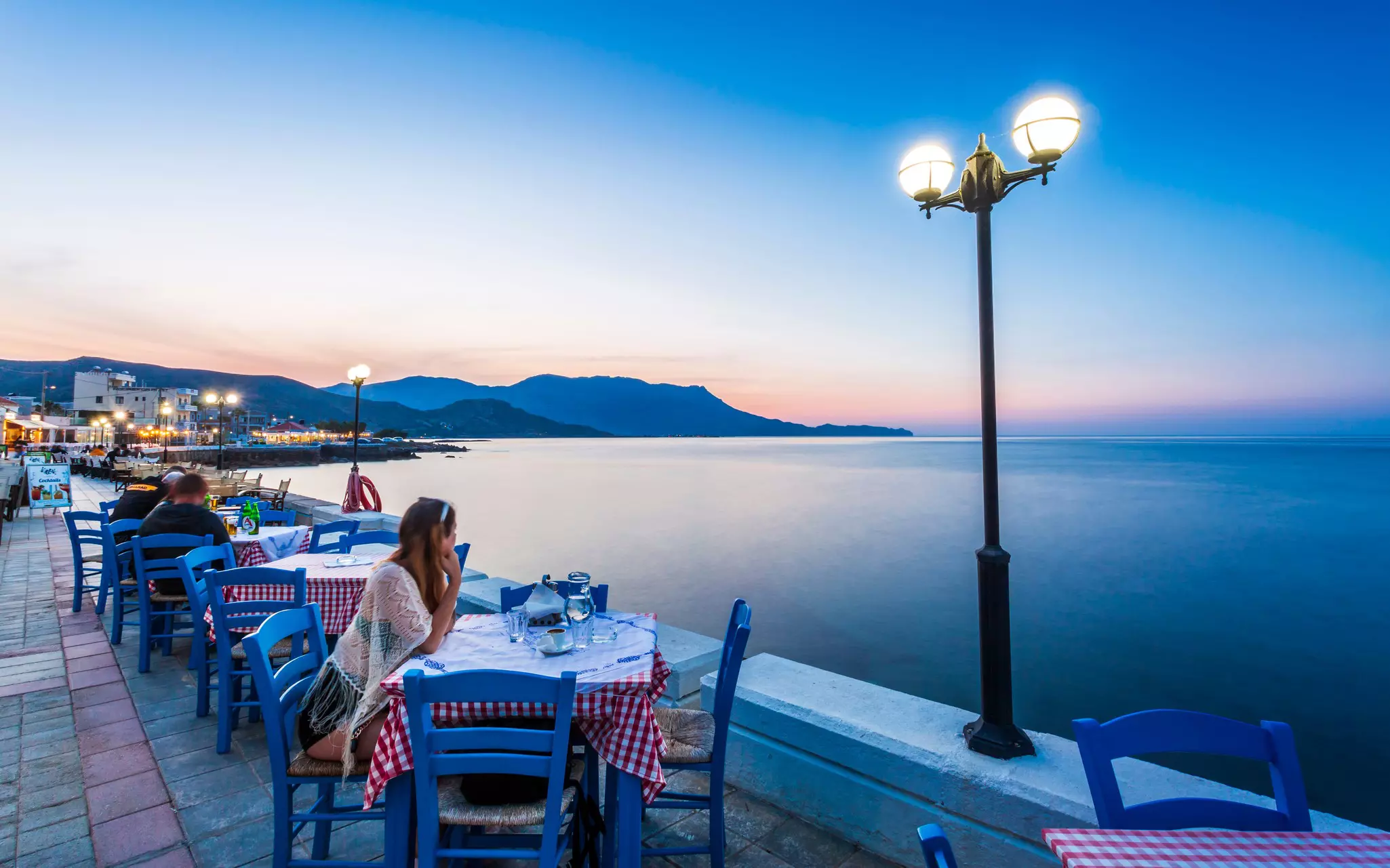 A women sits at a restaurant table on a waterfront gazing out to sea as the sun sets.